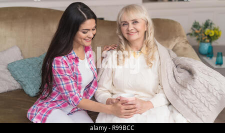 Lächelnd Tochter sanft berührt Ihr die Mutter die Hände. Elegante ältere Frau sitzt auf der Couch und hält eine Tasse Tee, während ihre junge Tochter umarmt sie. Stockfoto