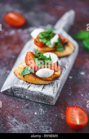 Italienische Bruschetta mit Tomaten, Mozzarella und Kräuter auf einem Holzbrett auf einem dunklen Hintergrund. Stockfoto