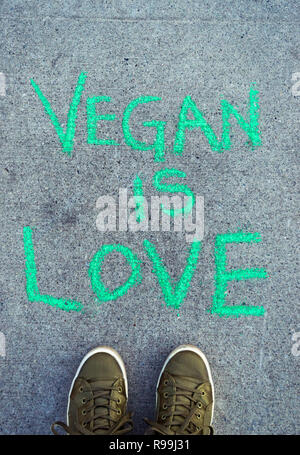 Text Wörter Vegan ist Liebe in Grüne Kreide Schriftzug auf Zement Bürgersteig mit POV grün Freizeitschuhe. Stockfoto