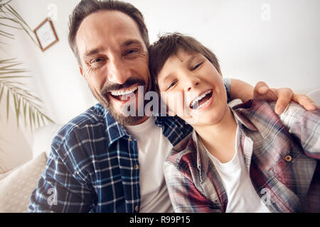 Vater und Sohn zusammen zu Hause sitzen auf einem Sofa umarmen unter selfie Fotos auf dem Smartphone suchen Kamera lacht fröhlich close-up Stockfoto