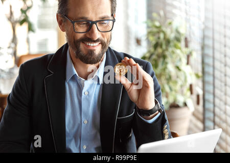 Reifer mann Händler Tragen von Brillen am Tisch im Cafe sitzen mit Laptop auf cryptocurrency Münze lächelt glücklich Stockfoto