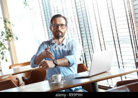 Reifer mann Händler Tragen von Brillen am Tisch im Cafe tagsüber sitzt mit Laptop und eine Tasse Kaffee in der Zeit zeigend auf Watch suchen Kamera Konzert Stockfoto