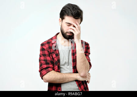 Xhausted junger Mann das Gesicht mit der Hand während gegen grauen Hintergrund stehend Stockfoto