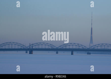 Eisenbahnbrücke und Fernsehturm im Winter. Eisenbahnbrücke über den zugefrorenen Fluss Daugava in Riga. TV Tower und Eisenbahnbrücke auf blauen Himmel Hintergrund Stockfoto