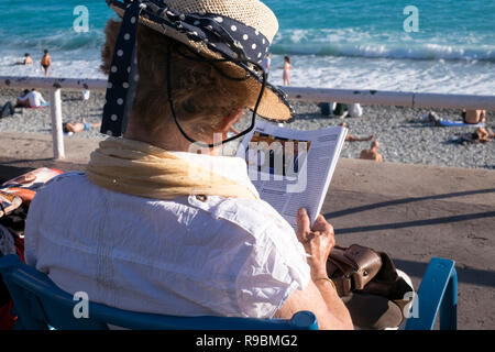 Eine ältere Dame in einem Strohhut sitzt am Meer das Lesen einer Zeitschrift mit Blick auf den mediterranen Meer. Stockfoto