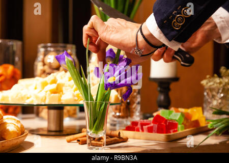 Hand des eleganten Mann mit Leder Armband schneidet Blumen von Messer, lila Iris in Glas Vase gegen ein Buffet mit Leckereien und Snacks. Konzept f Stockfoto