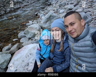 Touristen Rest nach der Wanderung und selfie am Telefon. Fotos vom Handy während der Wanderung. Familie reist. Menschen Umwelt durch Berge, Flüsse, Ströme. Mann und Frau haben wandern Rucksäcke. Stockfoto