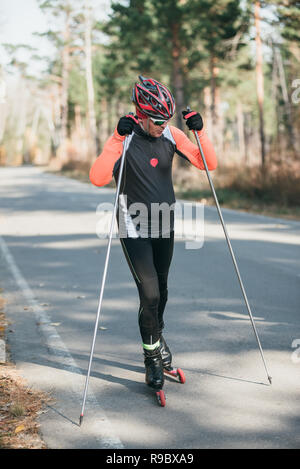 Training ein Athlet auf der Walze Skater. Biathlon Fahrt auf dem Roller Ski mit Skistöcke, im Helm. Athlet ist immer bereit zu starten. Herbst Training. Roller Sport. Stockfoto