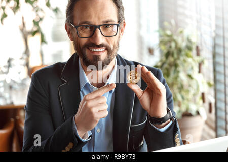 Reifer mann Händler Tragen von Brillen am Tisch im Cafe sitzen mit Laptop auf cryptocurrency Münze suchen Kamera lächelnd toothy Stockfoto