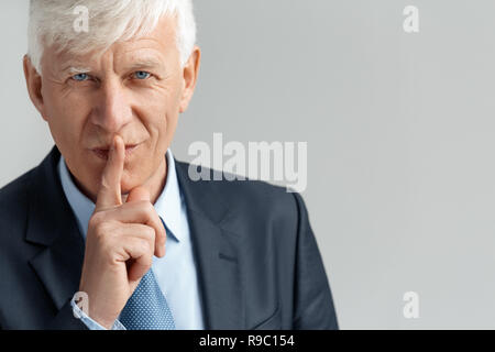 Senior Business mann Studio stehen isoliert auf graue Wand, ruhigen Geste geheim zu halten, die Kamera lächelnd verspielten close-up Stockfoto