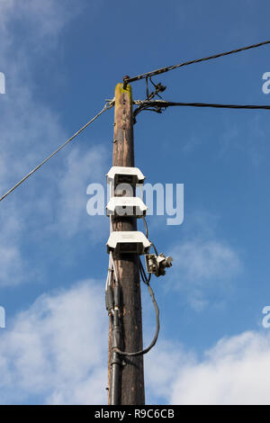 Eine hölzerne Stange unterstützt zwei schwarze Stromkabel an seiner Oberseite. Unten auf der Pole sind drei weiße Sicherungskästen. Der Himmel ist blau mit whispy weißen Wolken. Stockfoto