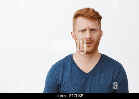 Redhead erwachsenen Mann in Erstarrung nicht verstehen, was geschieht, Gefühl, keine Ahnung und befragt, Augenbraue anheben und an der Kamera starrt verwirrt, unsicher über grauer Hintergrund Stockfoto