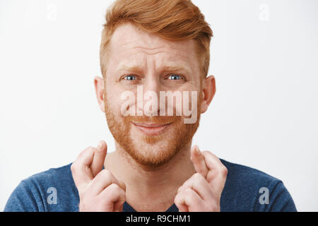 Studio geschossen von besorgt Intensive redhead miserablen Kerl mit Bart in blauen T-Shirt die Daumen, schürzte die Lippen und runzelte die Stirn, während nervös und Betteln für Glück und Erfüllung von Wünschen Stockfoto