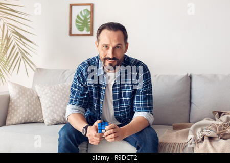 Der Mensch allein zu Hause sitzen auf einem Sofa holding Rubik's Cube suchen Kamera lächelt Fröhlich Stockfoto