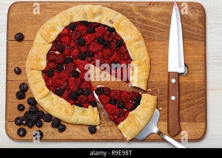 Hausgemachte Himbeeren Galette mit einem Stück weg auf einem hölzernen Schneidebrett schneiden. In der Nähe liegt das Messer und zerstreute Beeren. Ansicht von oben. Stockfoto