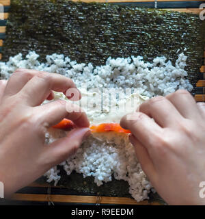 Die Sushi Rollen zu Hause: weibliche Hände Lachs Fisch Streifen auf Frischkäse, Reis, Nori Algen Blatt auf einem Bambus Matte gelegt. Ansicht von oben Stockfoto