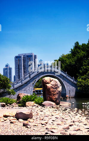 Eine gewölbte Brücke über einem See in: Lizhi Park in Futian District Shenzhen China mit der Skyline im Hintergrund. Stockfoto