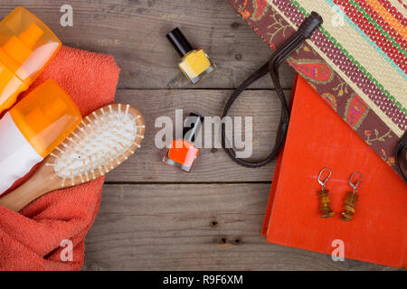 Strand Zubehör - Haarbürste, orange Handtuch, Sun Creme, Lotion, Strandtasche, Nagellack, ein Buch auf einem braunen Holz- Hintergrund Stockfoto