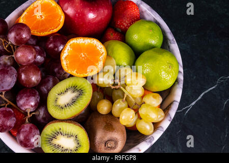 Frische tropische fruitson in Schüssel auf schwarzem Marmor Hintergrund. Ananas, Papaya, Mango, Kiwi, Trauben, Granat, Mandarinen, Bananen, Äpfel, Wassermelone sli Stockfoto