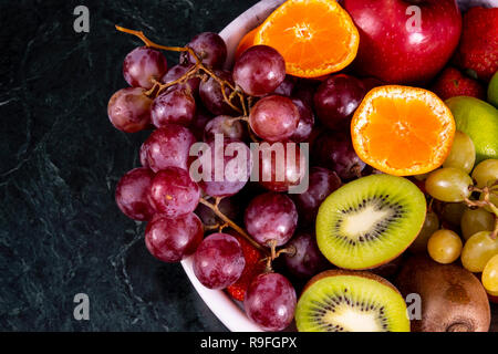 Frische tropische fruitson in Schüssel auf schwarzem Marmor Hintergrund. Ananas, Papaya, Mango, Kiwi, Trauben, Granat, Mandarinen, Bananen, Äpfel, Wassermelone sli Stockfoto