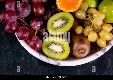 Frische tropische fruitson in Schüssel auf schwarzem Marmor Hintergrund. Ananas, Papaya, Mango, Kiwi, Trauben, Granat, Mandarinen, Bananen, Äpfel, Wassermelone sli Stockfoto