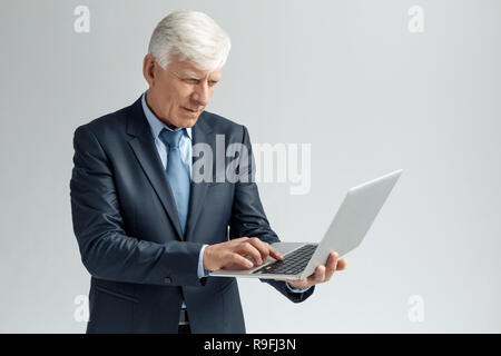 Senior Business mann Studio stehen auf graue Wand holding Laptop surfen Website isoliert konzentriert Stockfoto