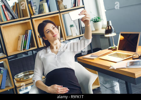 Schwangere Frauen arbeiten im Büro drinnen sitzen berühren Bauch posing unter selfie Fotos für soziale Medien freudige Stockfoto
