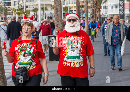 Benidorm, Costa Blanca, Spanien, 25. Dezember 2018. Britische Touristen Kleid für den Anlass am Weihnachtstag in diesem beliebten Ausflugsziel für Briten entkommen dem kalten Wetter zu Hause. Die Temperaturen werden in der Mitte der heute zu hohen 20 Celsius in diesem mediterranen Hotspot. Paar tragen Weihnachten Kleidung und Nikolausmützen. Paar mittleren Alters tragen leuchtend roten Weihnachten Jumper und Hüte, mit Santa Bart waliking draußen in der Sonne. Stockfoto