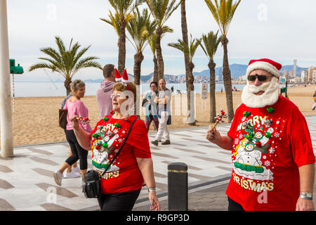 Benidorm, Costa Blanca, Spanien, 25. Dezember 2018. Britische Touristen Kleid für den Anlass am Weihnachtstag in diesem beliebten Ausflugsziel für Briten entkommen dem kalten Wetter zu Hause. Die Temperaturen werden in der Mitte der heute zu hohen 20 Celsius in diesem mediterranen Hotspot. Paar tragen Weihnachten Kleidung und Nikolausmützen. Paar mittleren Alters tragen leuchtend roten Weihnachten Jumper und Hüte, mit Santa Bart waliking draußen in der Sonne. Stockfoto