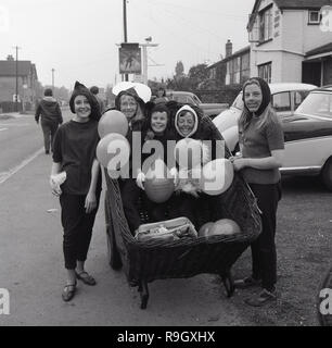 1967, prestwood Dorffest, einer Gruppe von Mädchen Spaß... gekleidet sind Sie draußen auf der Straße stehen, drei von ihnen in einem großen Weidenkorb auf einer Karre holding Ballons, Buckinghamshire, England, Großbritannien sitzen. Stockfoto