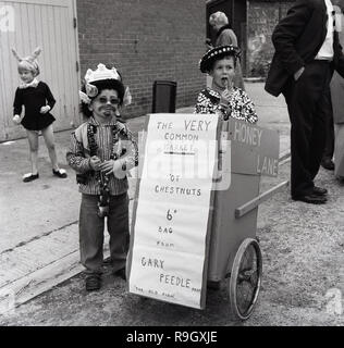 1967, historischen, zwei Jungen in Kostüm Kostüm - Eine als East End von London Pearly King - Verkauf von Kastanien in 6D eine Tasche aus ihren kleinen hausgemachten Warenkorb in der Straße am Prestwood Dorffest. Stockfoto