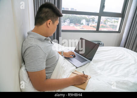 Mann mit Laptop und Schreiben Notizen im Schlafzimmer Stockfoto