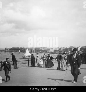 Alate Viktorianischen schwarz-weiß Foto im Englischen Badeort von Morecambe, Lancashire, England. Es zeigt einen Mann mit schwarzer afrikanischer Abstammung, in feinen Kleidern bekleidet und mit einem Spazierstock, der Promenade hinunter, während ein kleiner Junge schaut ihn an. Es gibt noch viele andere Leute im Hintergrund, die die Art und Weise der Tag, sowie kleine Boote im Hafen. Stockfoto