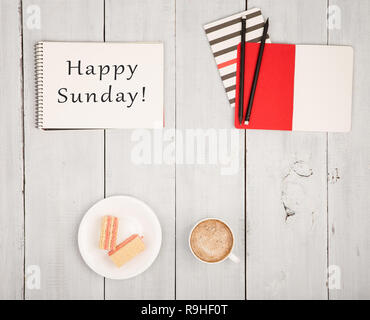 Tabelle mit Notizblöcken und Text "Happy Sonntag!', Tasse Kaffee und Waffeln auf weißem Holz- Hintergrund Stockfoto