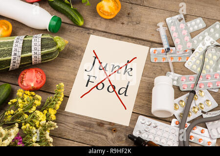 Die Wahl zwischen einem gesunden Lebensstil und Medikamente. Gemüse oder Pillen. Papier mit Text "kein Junk Food', Stethoskop auf braunem Holz- Schreibtisch Stockfoto