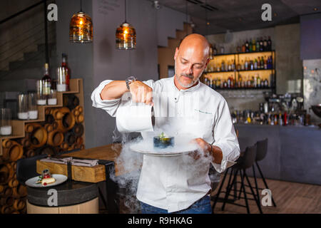 Schönen italienischen Mann mit Glatze mit Bart in weiße Uniform Koch in einem Restaurant mit einem toothy Lächeln in seinen Händen eine Platte von Dessert posieren. Stockfoto
