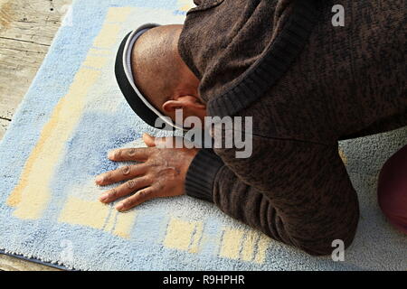 Ein muslimischer Mann betet mit einer Matte auf dem Boden und einem Foto von Menschen Stockfoto