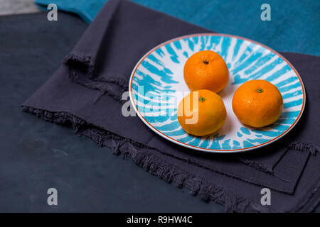 Drei reife lecker saftig Mandarin liegen in einem hellen Weiß mit einem blauen Platte Muster auf dem Stoff tuch Küchentuch auf einem dunkelblauen schwarzen Hintergrund. Stockfoto