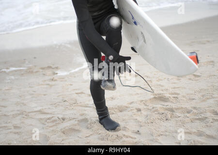Nahaufnahme Porträt einer Surfer mit dem Surfbrett auf dem Hintergrund von Sand und Meer. Kerl in einen Taucheranzug korrigiert. Surfschule Stockfoto