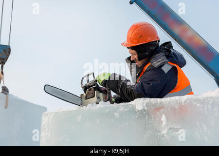 Workman Assembler in einem Schutzhelm mit einer Kettensäge in der Hand schneidet den Eisblock Stockfoto