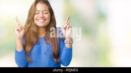 Junge schöne Brünette Frau trägt blaue Pullover über isolierte Hintergrund lächelnd die Daumen mit Hoffnung und die Augen geschlossen. Glück und superstitiou Stockfoto