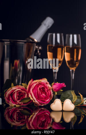 Flasche Champagner im Eimer mit zwei Weingläser, Blumenstrauß aus Rosen, herzförmige Pralinen und Kerzen auf schwarzen Hintergrund. Liebe, Valentines Tag c Stockfoto