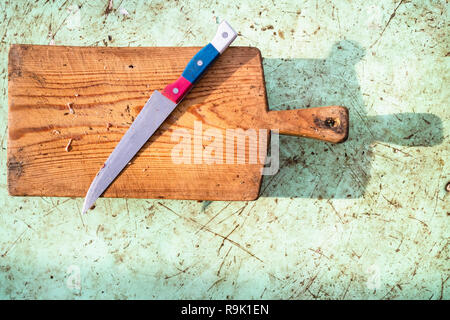 Ansicht von oben aus Stahl Messer mit einer rot-blau-weißer Griff auf einen schmutzigen Holz Schneidebrett auf einem Outdoor unordentlich Tabelle Stockfoto