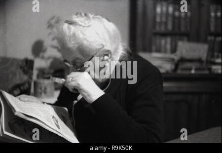 1950er Jahre, ältere Dame in einem Zimmer vorne mit einem Handheld Lupe die Drucken bis zu schließen Auf ein Magazin, England, UK lesen Sie sitzen. Die Objektiv montiert in einem Frame hilft Vergrößern Vergrößern oder Drucken in einer Zeitung oder einer Veröffentlichung dabei helfen denen mit schlechten oder fehlerhaften Sehvermögen. Stockfoto