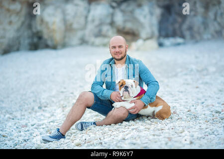 Stattliche bold Stilvolle in Jeans Mann in blaue Kappe am Strand Meer sitzen zusammen mit hübschen englischen Bulldogge gekleidet Stockfoto