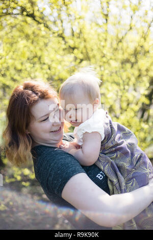 Mutter lächelnd, während sie ihr Baby wickeln in einer Schlinge für babywearing. Lifestyle Bild geschossen auf Lage in einem Park an einem sonnigen Frühlingstag. Stockfoto