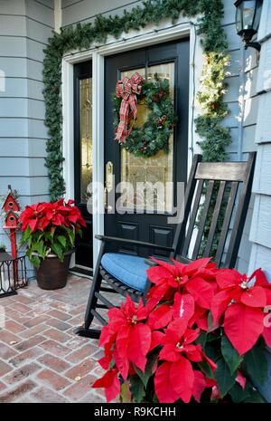 Vordere Tür Eingangsbereich mit traditionellen amerikanischen Weihnachten Dekor: poinsettias, Kranz, Laternen, rot Vogelhaus, Evergreens und einem Schaukelstuhl. Stockfoto