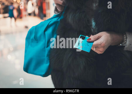 Gut gekleidete Dame im Pelz holding Kreditkarte und Tasche auf unscharfen Mall Hintergrund. Frau Zahlung in der Mode Boutique, Bankkarte zu Kasse. Weiblich Stockfoto