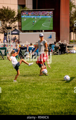 Während tagsüber ein Publikum Uhren eine Fußball-WM Spiel auf einen gigantischen Outdoor TV-Bildschirm in Costa Mesa, CA, eine Mutter mit ihren Kindern das Spiel auf einem nahe gelegenen Feld spielt. Stockfoto