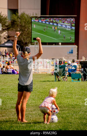 Während tagsüber ein Publikum Uhren eine Fußball-WM Spiel auf einen gigantischen Outdoor TV-Bildschirm in Costa Mesa, CA, eine Mutter mit ihren Kindern das Spiel auf einem nahe gelegenen Feld spielt. Stockfoto